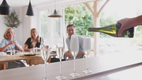 Ein-älterer-Mann-öffnet-Eine-Flasche-Champagner,-Während-Die-Familie-Mit-Erwachsenen-Nachkommen-Zu-Hause-Am-Tisch-Isst