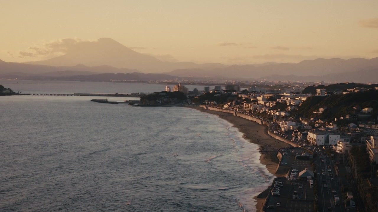 Costa di Shichirigahama a Kamakura, strada costiera ed edifici collinari che si estendono verso l'isola di Enoshima, con la sagoma sfocata del Monte Fuji che domina l'orizzonte sotto la calda luce del tramonto.