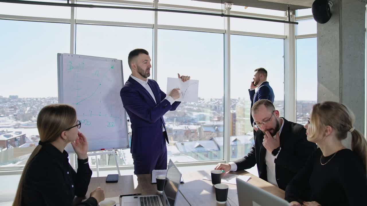 Businessman holding a paper with graph on it and showing it to his colleagues. Coworkers sitting at the table and looking at the paper. Man in a suit standing at the window and talking on the phone at the backdrop.