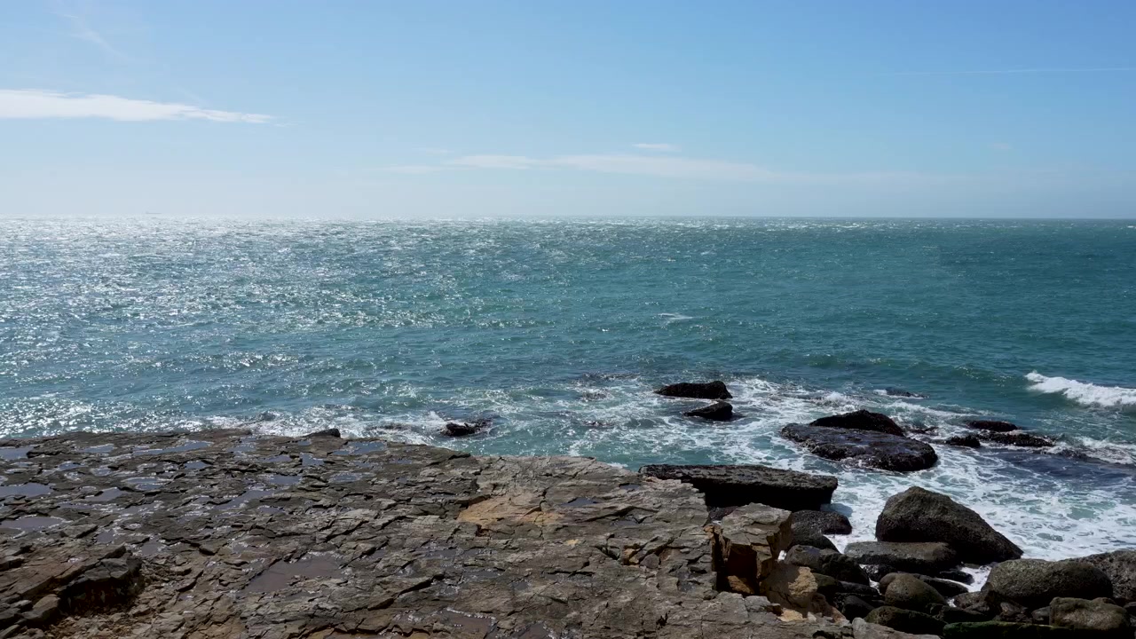 Plano geral cinematográfico de água azul do mar atingindo uma costa rochosa acidentada sob um céu limpo. Adequado para conteúdo de viagens e natureza
