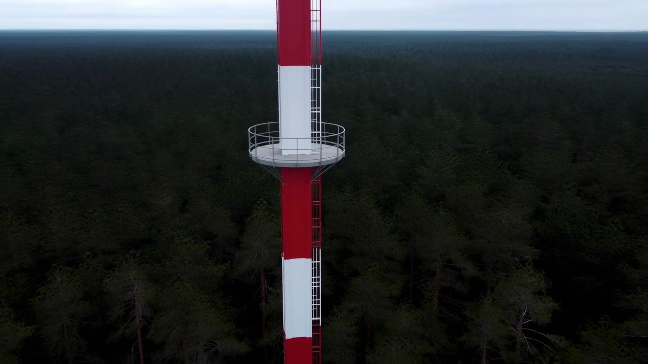 Centered aerial view of circular metal observation platform fixed to tall red and white tower above continuous evergreen forest, showing industrial structure, safety railing, vertical form, remote