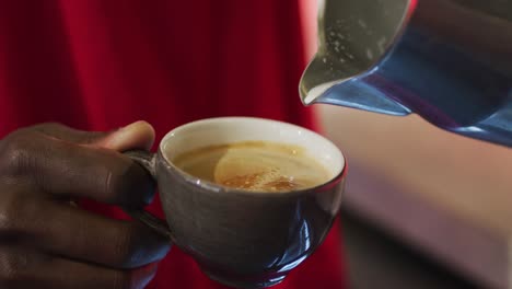 midsection of african american barista pouring milk from jar into coffee cup in cafe