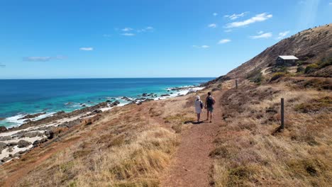 couple walking along a remote dirt path with a small shack on their right and the bright blue ocean and white sand beach to their left