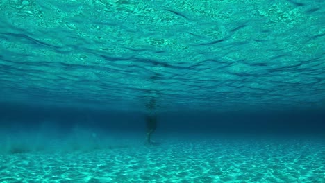 Beautiful-underwater-scene-of-man-walking-on-sandy-sea-floor-with-sunlight-refractions-and-reflections-in-turquoise-transparent-ocean-water