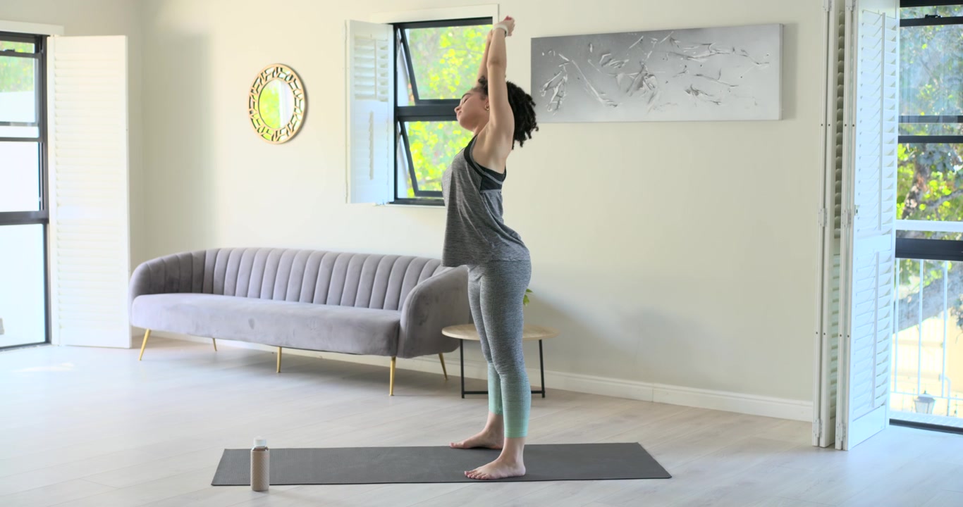 Mulher afro-americana em pé sobre um tapete de yoga, alongando-se, torcendo-se e dobrando-se para se esticar em casa.