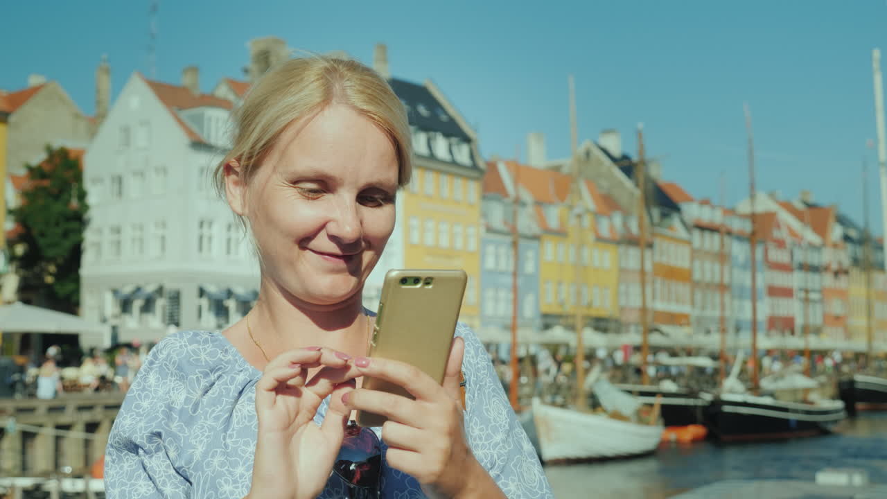 행복한 여성은 스마트폰을 사용하여 운하와 인기 있는 관광 거리의 배경에 서 있습니다.