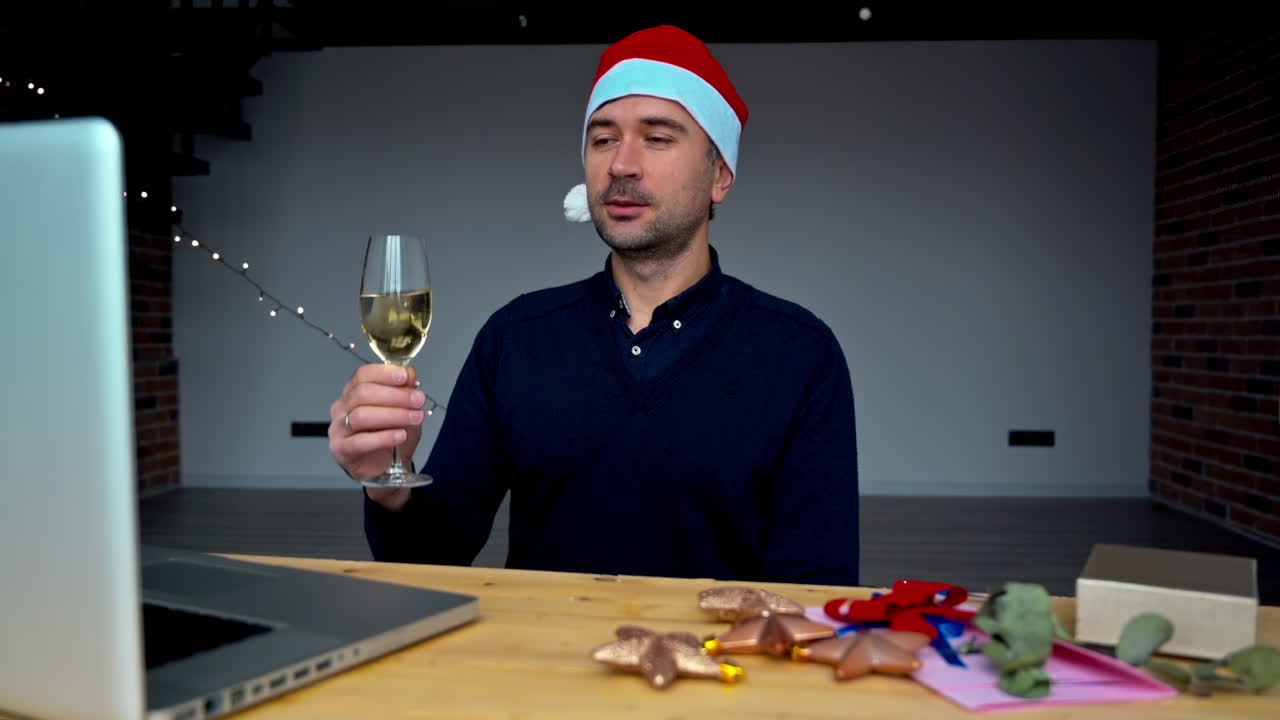 A man wearing a festive hat raises a glass for a toast in a cozy workspace decorated for the holidays. Cheerful decorations and gifts surround him as he enjoys a moment of celebration
