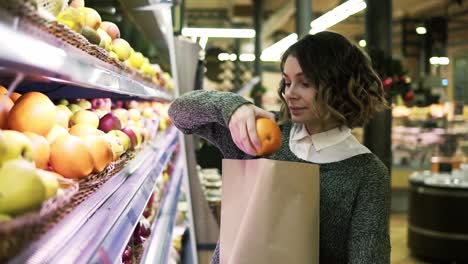 cute girl buys fresh oranges in the market. beautiful young woman stands in front the shelf and puts the oranges to a brown