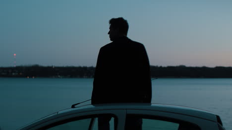 Young Caucasian male sitting on car roof at blue hour by the sea