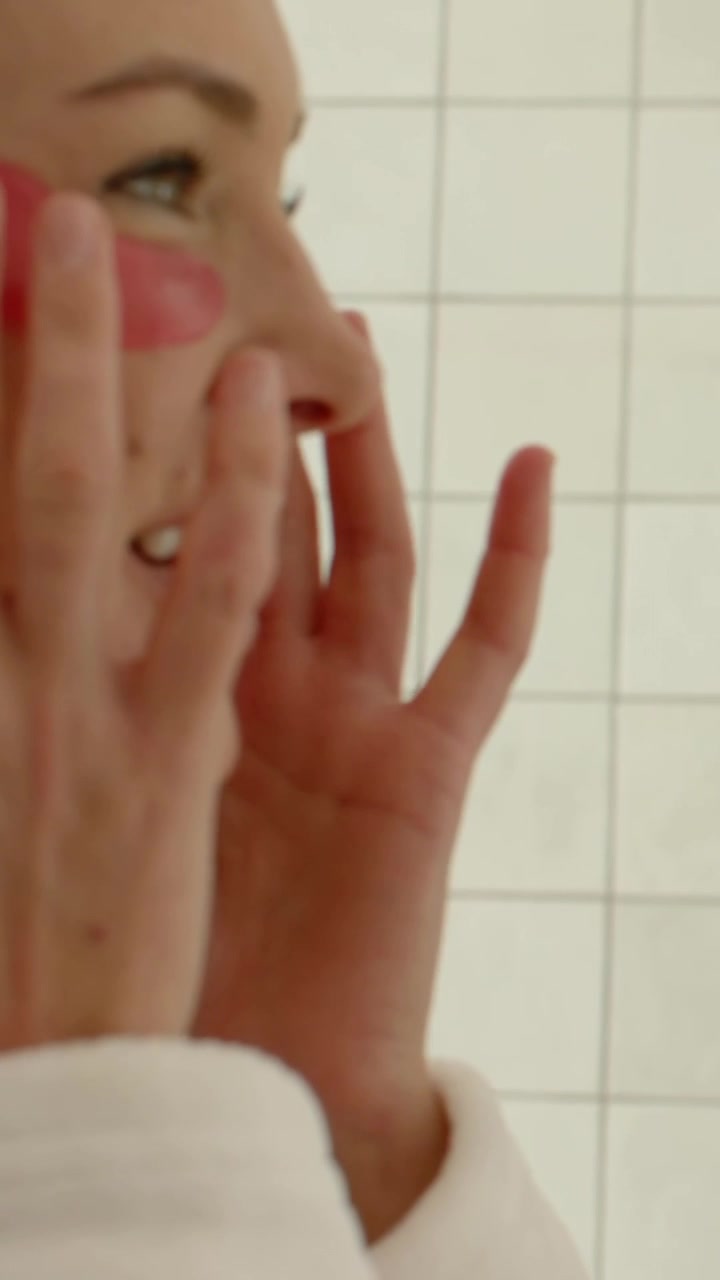 Video verticale: Donna in accappatoio bianco che applica una patch rossa sotto gli occhi, stendendola sulla guancia davanti a una parete piastrellata.
