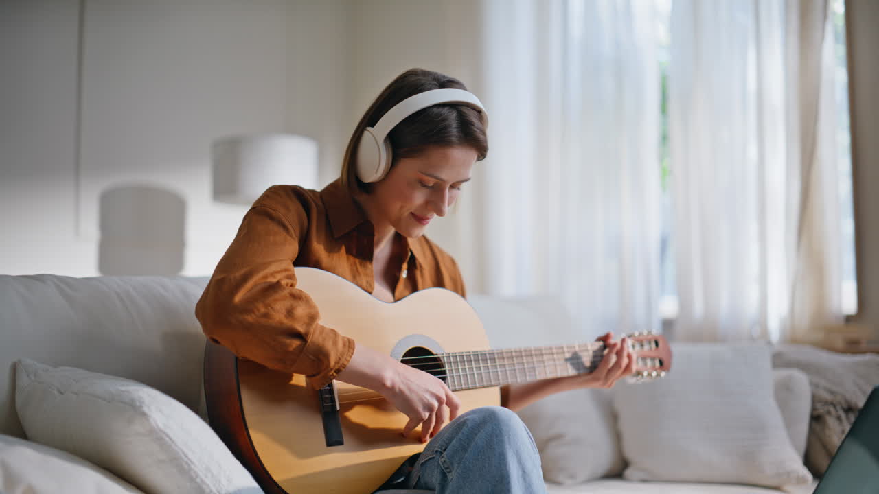 Smiling girl playing guitar in headphones watching tutorial laptop home closeup