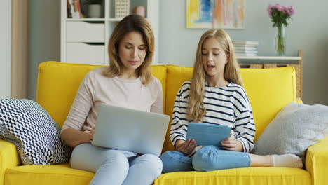 blonde mother and daughter sitting on yellow couch while they talking and using laptop computer and tablet in the living room