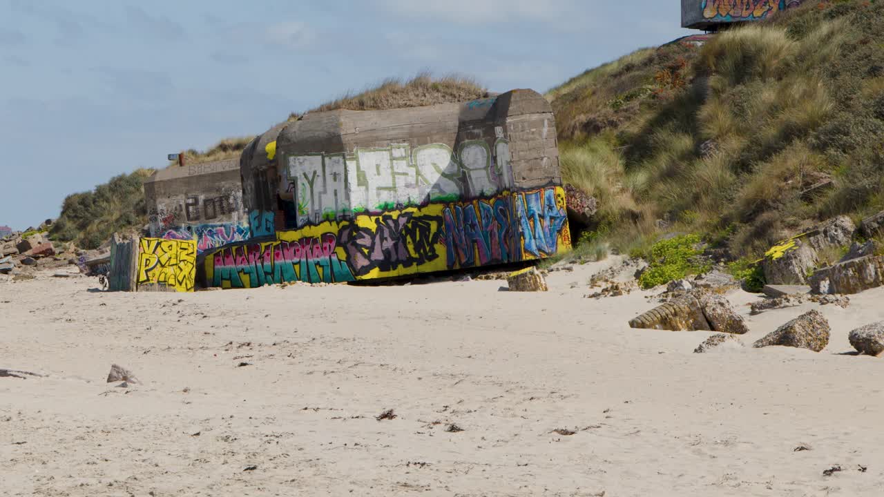 Daylight pan reveals graffiti-covered war bunker, sandy dunes, rocky debris, and distant coastline