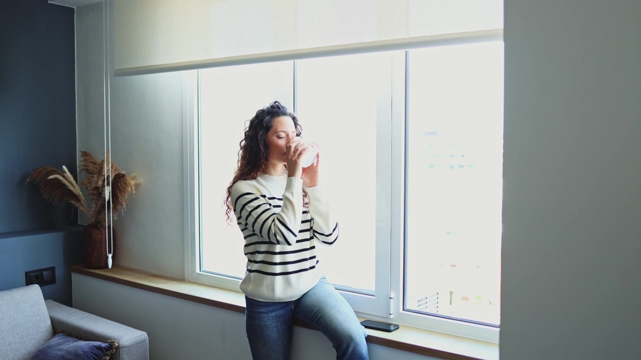Mujer joven usando un smartphone y bebiendo café junto a la ventana