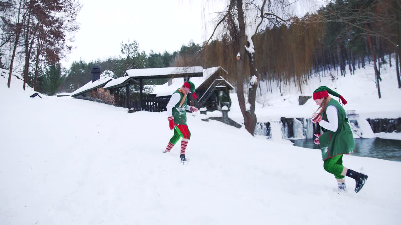 Elves in green costumes and long hoods play in snow. Winter background and two happy fairy characters playing snowballs before the house in the forest
