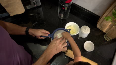 Foto-Inclinada-Hacia-Abajo-De-Un-Padre-Mexicano-Irreconocible-Con-Su-Hijo-Tamizando-Harina-Golpeando-Un-Colador-En-La-Cocina