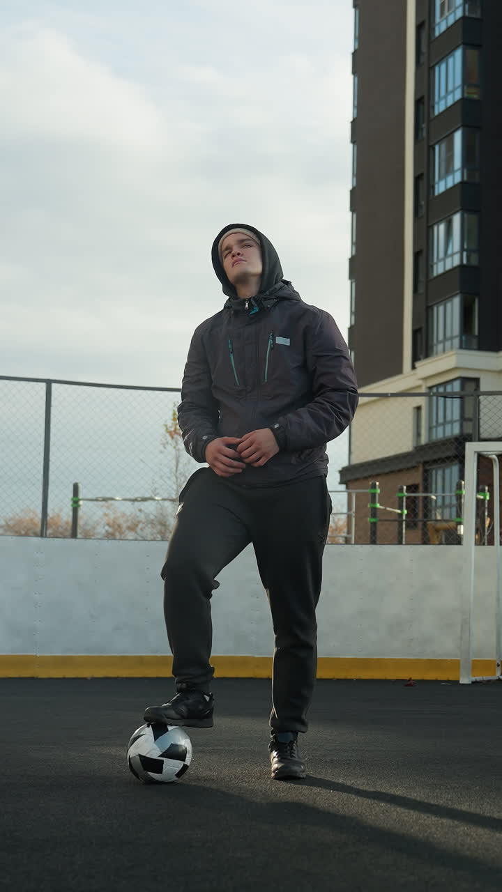 Athlete wearing hoodie with leg on ball stretching arms wide, turning head in urban sports arena, surrounded by goalpost and tall residential buildings