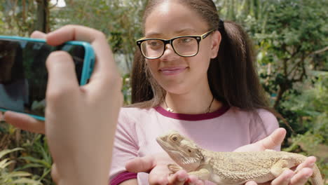 chica valiente sosteniendo una lagartija iguana con un amigo tomando fotos usando un teléfono inteligente compartiendo una excursión al zoológico en las redes sociales aprendiendo sobre reptiles en el santuario de vida silvestre 4k