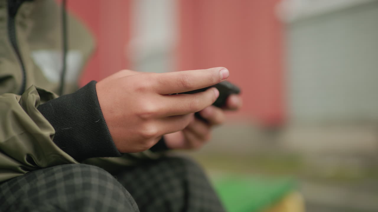 Close up of kid hands holding handheld device while playing game outdoors, dressed in green jacket with blurred bokeh background of urban environment creating casual lifestyle scene