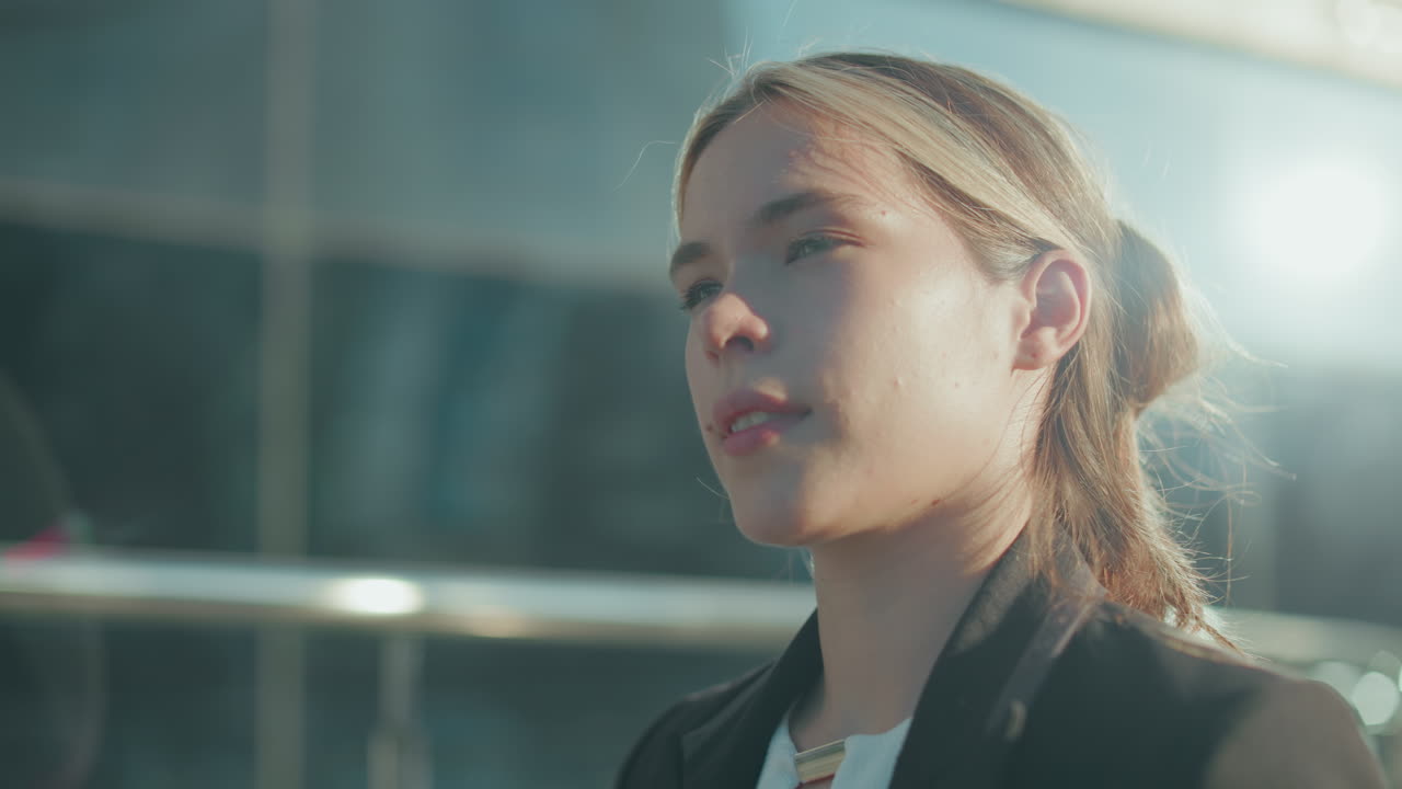 Side view of young lady walking confidently while holding file folder and coffee cup, sunlight illuminating her face, with metal railing and glass building reflecting ambient brightness in background