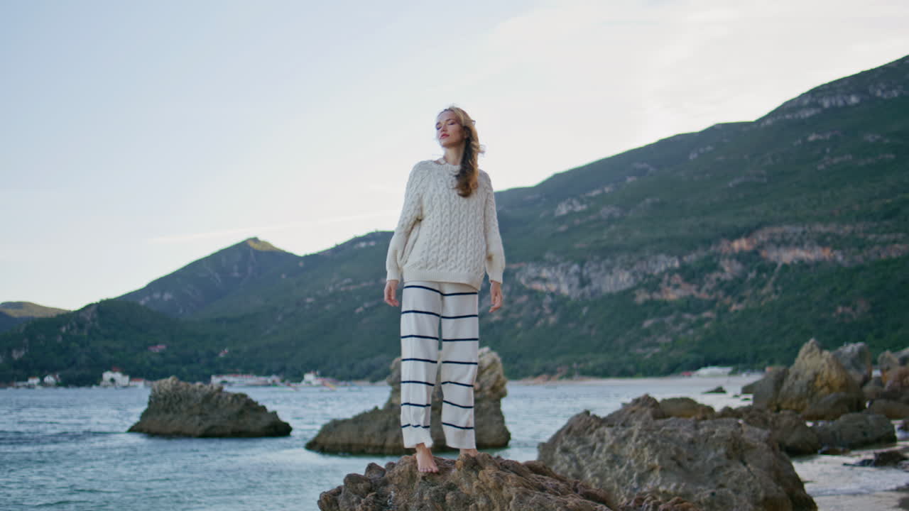 Frau genießt windigen Morgen am Meer, stehend auf Küstenfelsen. Entspannte Dame hebt die Hände