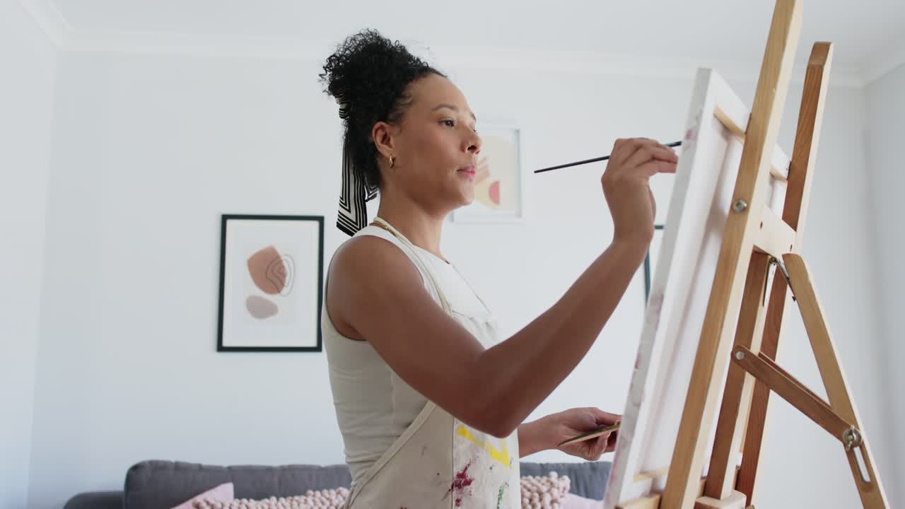 Donna afroamericana che carica il pennello dalla tavolozza, crea arte su tela su cavalletto in studio, con grembiule