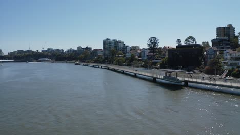 Toma-Amplia-De-Un-Dron-Volando-A-Lo-Largo-Del-Río-Brisbane-En-New-Farm,-Siguiendo-A-Un-Grupo-De-Ciclistas-Mientras-Recorren-El-New-Farm-River-Walk.