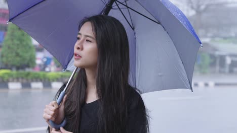 young asian female with a long black hair disappointed stuck in heavy rain outside, hand using umbrella to prevent getting wet, facial expression of negative feeling, tropical climate changes