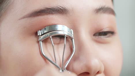 close up of beautiful young asian woman using an eyelashes curler while applying makeup at home