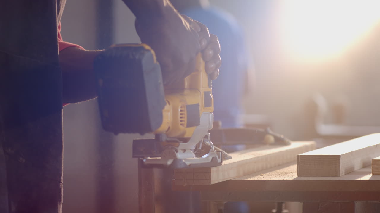 Cutting wooden plank with jigsaw in woodshop by African American male coworkers, copy space