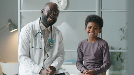Portrait-of-Cheerful-African-American-Doctor-and-Little-Boy-in-Clinic