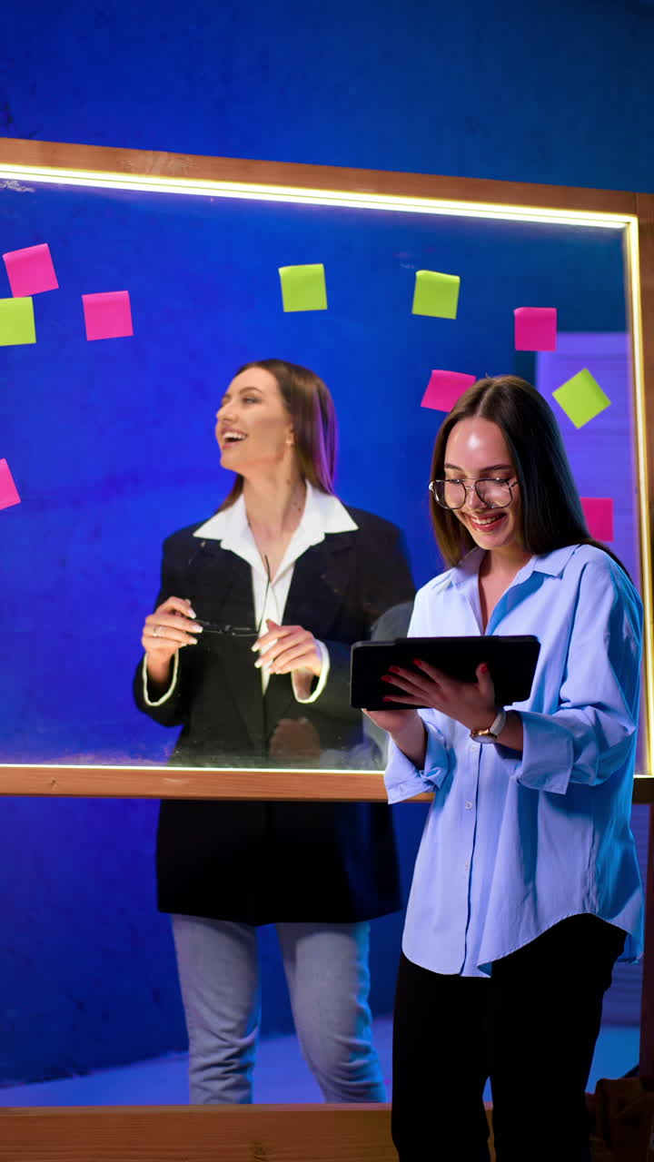 Positive female workmates stand at the glass wall. Ladies smile, talk, look at sticky notes. Producing ideas. Vertical video.