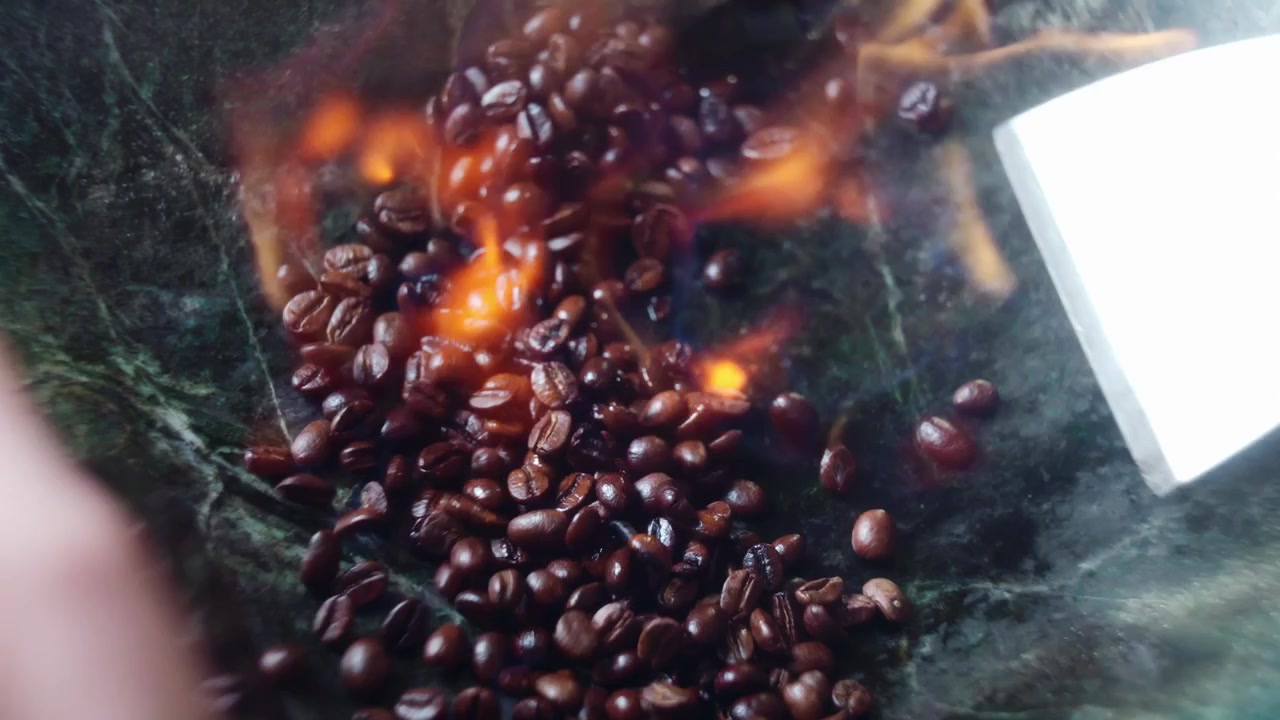 Primo piano della mano di un barista che usa una spatola d'acciaio per tostare chicchi di caffè su una superficie di pietra. Fuoco e liquore alle erbe accendono la miscela artigianale in un caffè boutique.