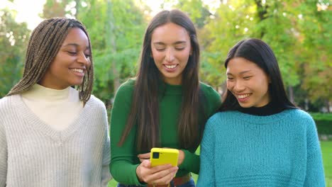 three diverse friends sharing moments and laughing with a smartphone in a park