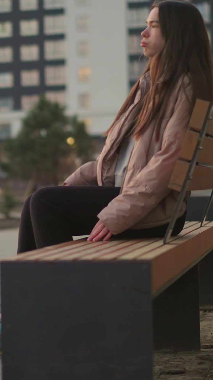A girl in a peach jacket and black trousers is seen rollerblading towards a park bench. The image captures the moment before she sits down to rest, with her rollerblades visible on the paved path