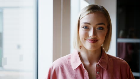 Young-white-woman-standing-by-the-window-in-an-office-turns-to-camera-and-smiles,-head-and-shoulders,-close-up