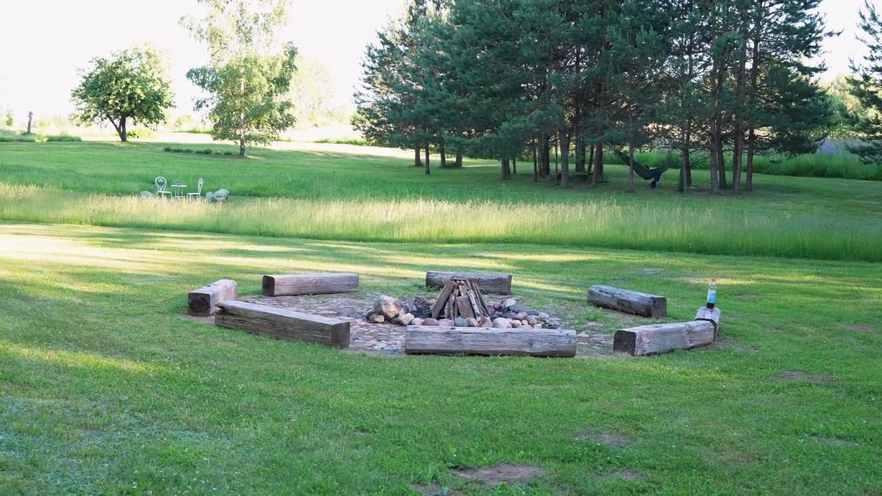 Lugn vidvinkelbild av en tom rustik eldstad omgiven av träbänkar på en ljusgrön äng, inramad av solljus, högt gräs och tallar – en perfekt fridfull sommarscen utomhus.