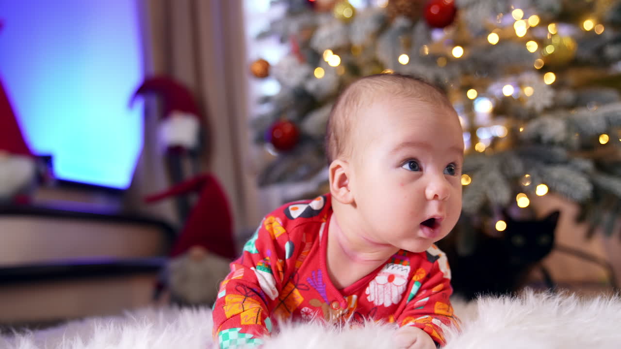 Infant baby in red costume lies on his belly in front of new year tree. Beautiful child tries to hold his head up and then falls to his side.