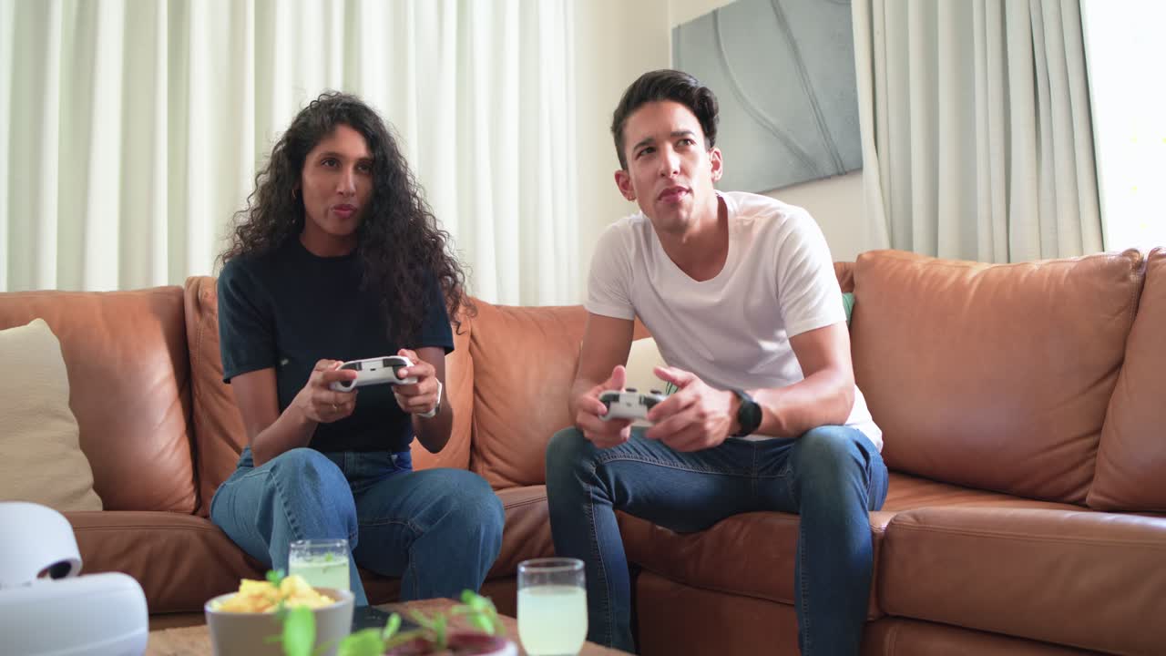 Amici afroamericani che reagiscono all'inizio di un gioco, con i controller bianchi in mano, seduti sul divano e mentre fanno uno spuntino.