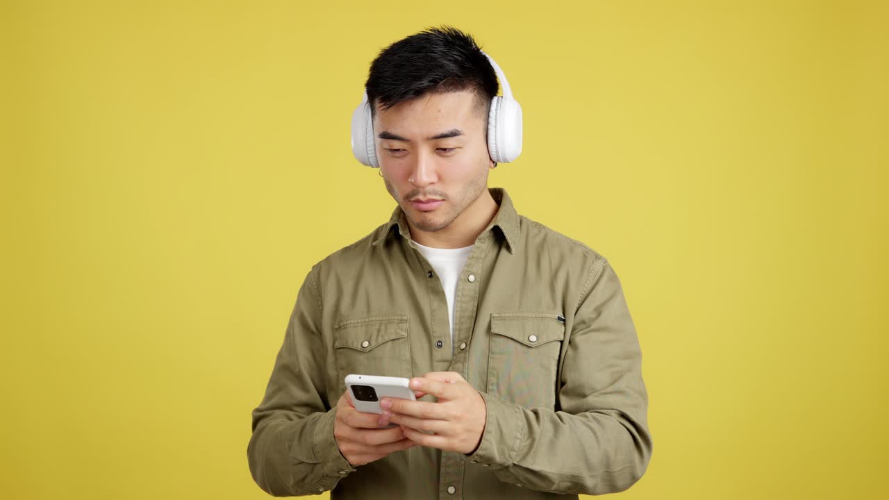 Young Asian man with headphones using smartphone on yellow background