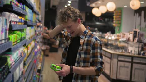 caucasian young man holding feminine pads in the mall and reading the label, grimacing. concept of shopping and choices. side view