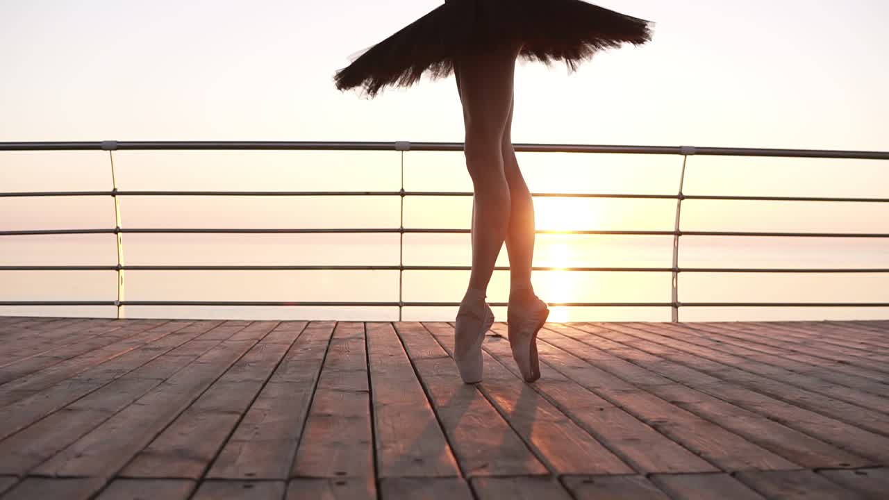 Close up of an elegant ballet dancer's legs, stepping on a wooden embankment on tip toes in pointes. Black ballet tutu. Beautiful scene with a morning sun rising in perspective