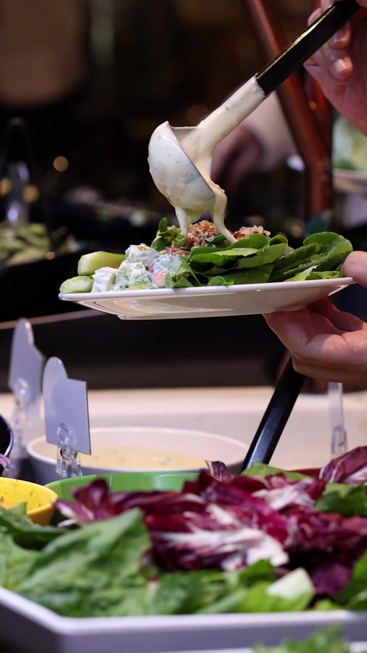 Primo piano di un commensale che versa condimento cremoso su insalata mista a un buffet di insalate self-service.