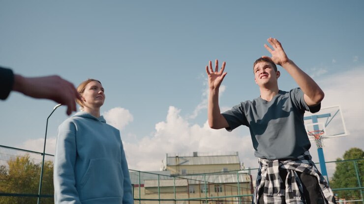 Sessione di addestramento di principianti che giocano a pallavolo con un allenatore con una camicia a quadri legata intorno alla vita in un campo aperto, sullo sfondo un edificio e una recinzione, che praticano tecniche di pallavolo