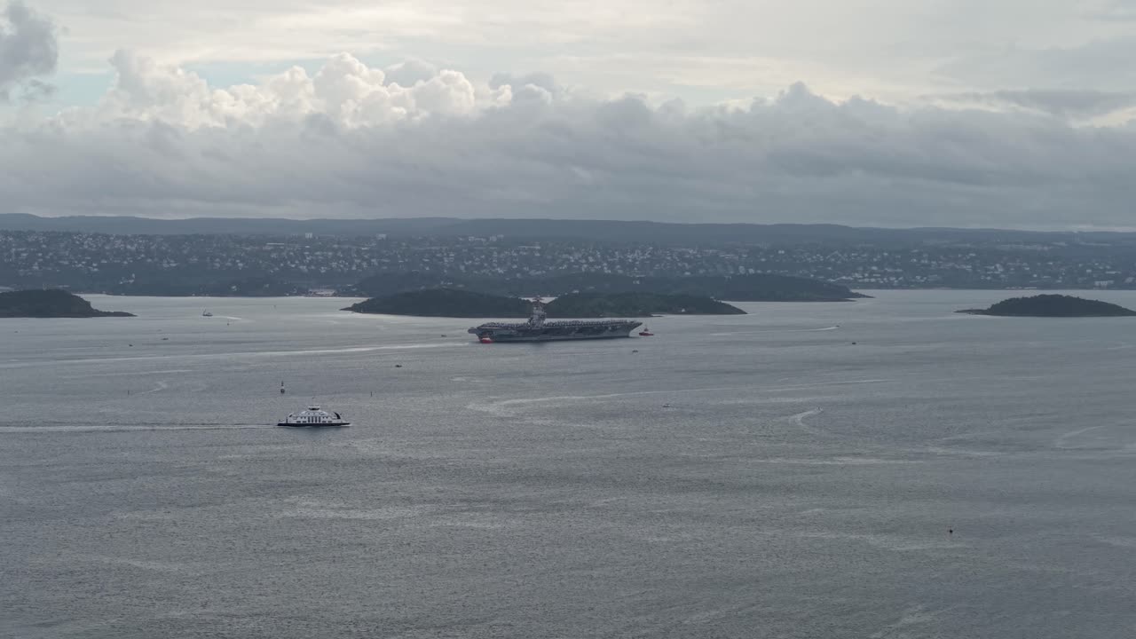 Boat Cruising In The Fjord Passing By The USS Gerald R. Ford Aircraft Carrier Ship In Oslo, Norway. - aerial shot