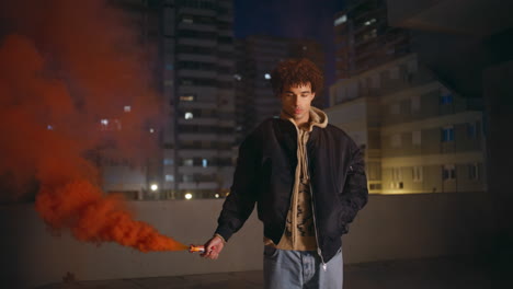 stylish guy smoke flare at nighttime urban setting. curly man holding grenade
