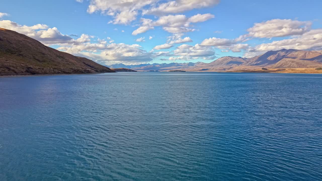 Aerial drone footage flying smoothly over Lake Tekapo in New Zealand, showing bright blue water, mountains and clouds under a clear sky