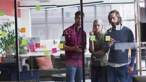 Diverse-office-colleagues-writing-on-glass-board-and-discussing-together-at-modern-office