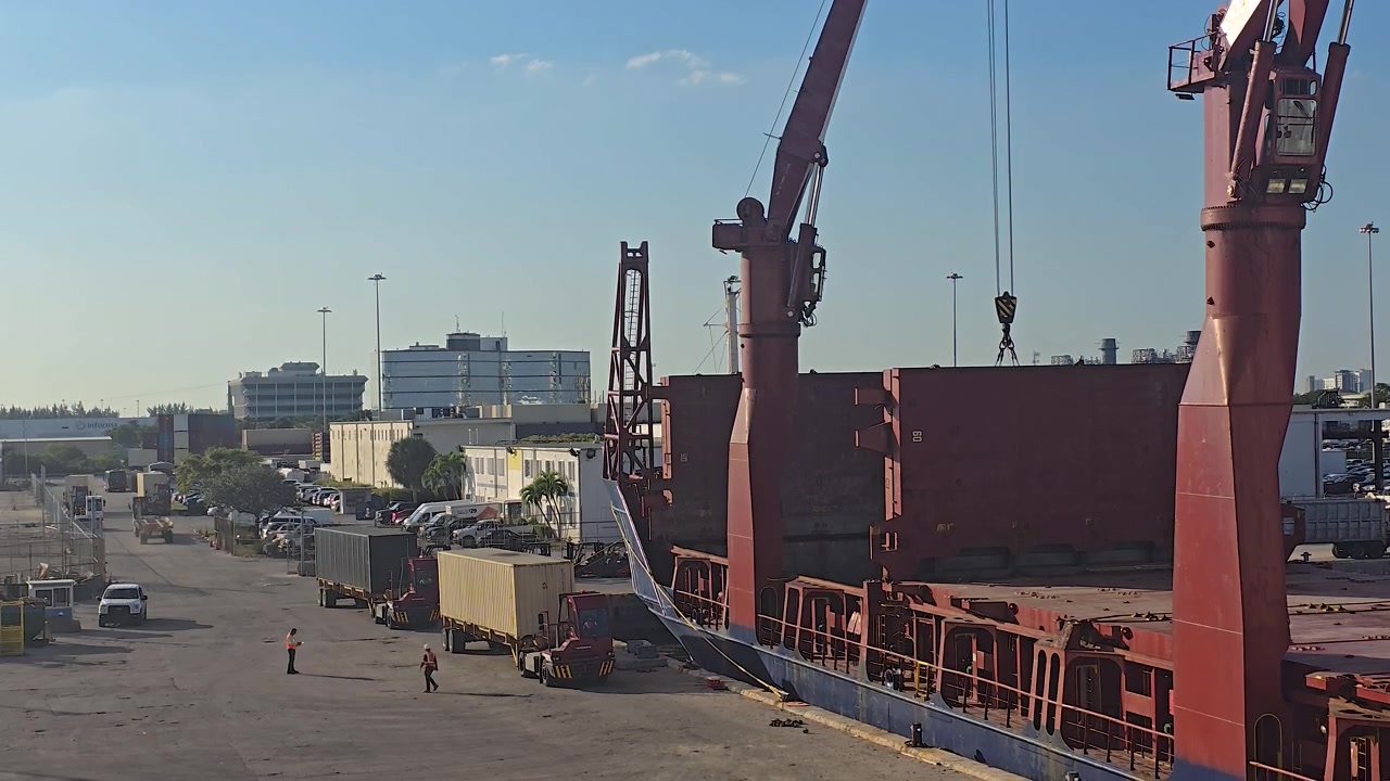 Una grande nave mercantile rossa attraccata in un trafficato porto industriale. Gru pesanti sono pronte per caricare merci mentre camion e lavoratori operano sul molo durante una giornata limpida.