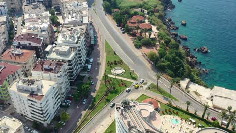 Drone-Aéreo-Que-Rodea-Una-Carretera-Costera-En-Antalya,-Turquía,-En-Un-Soleado-Día-De-Verano,-Mientras-Los-Autos-Salen-De-Un-Semáforo-Rodeado-De-Altos-Edificios-Residenciales